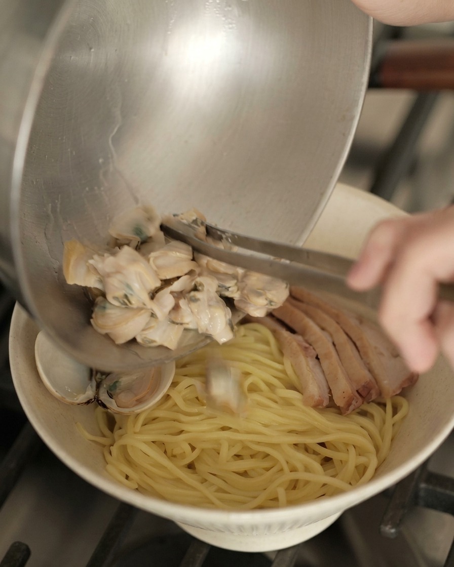 Vertiendo almejas cocinadas desde un cuenco metálico a un bol de ramen preparado con fideos y pato.