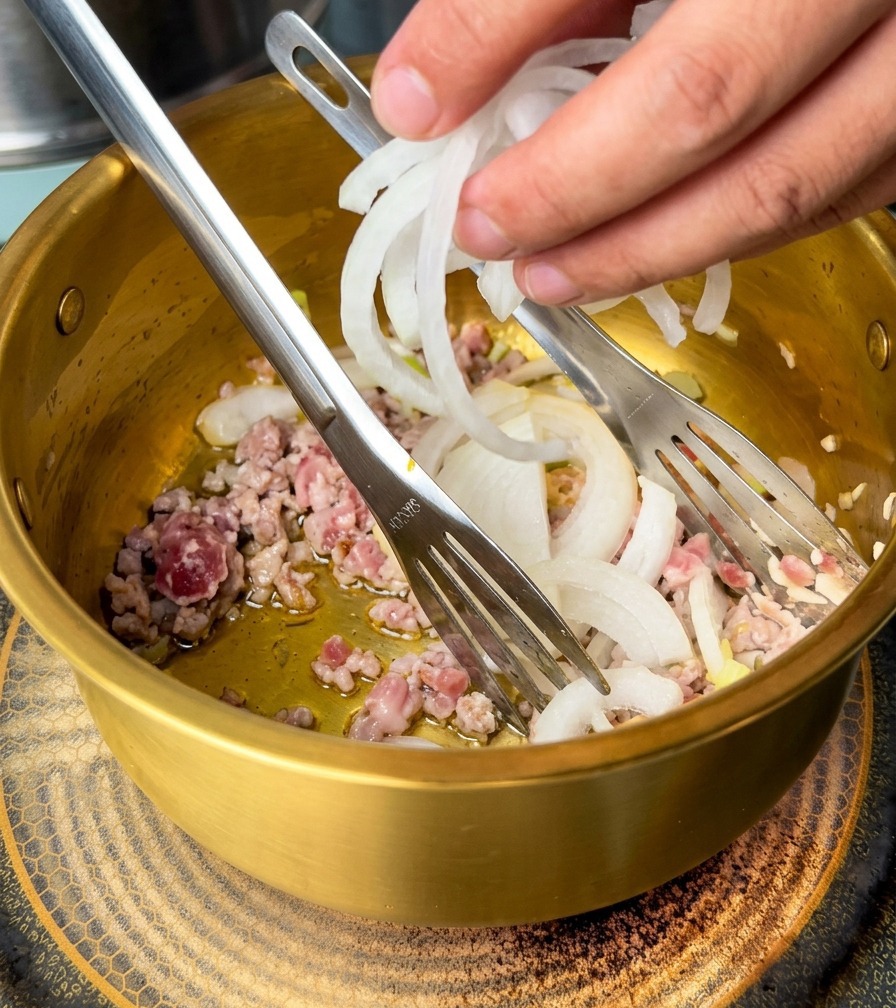 Mano añadiendo cebollas blancas cortadas a una olla dorada que contiene carne de cerdo y aromáticos.