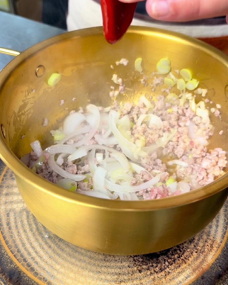 Salsa bibimbap roja vertiéndose en una olla dorada con cerdo y cebollas salteadas.