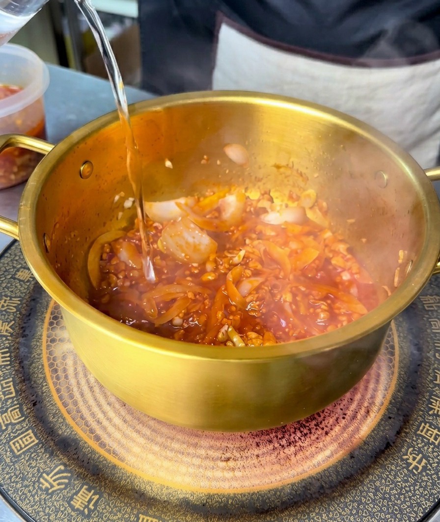 Agua clara vertiéndose en una olla dorada que contiene una base de carne y verduras roja y picante.