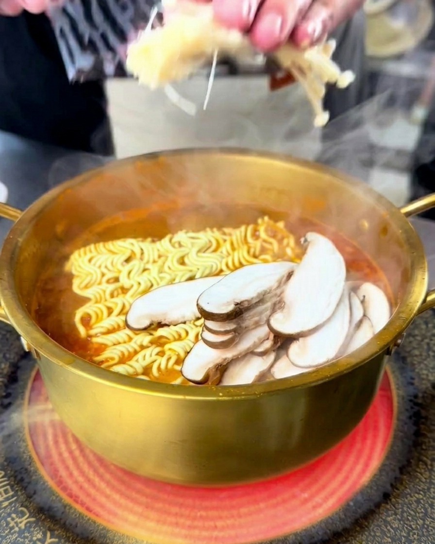 Persona añadiendo setas enoki y shiitake a una olla hirviendo de ramen de mariscos.