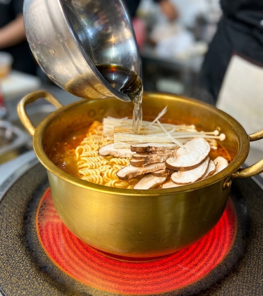 Añadiendo un cuenco de setas y mariscos a una olla dorada llena de fideos ramen y caldo rojo.