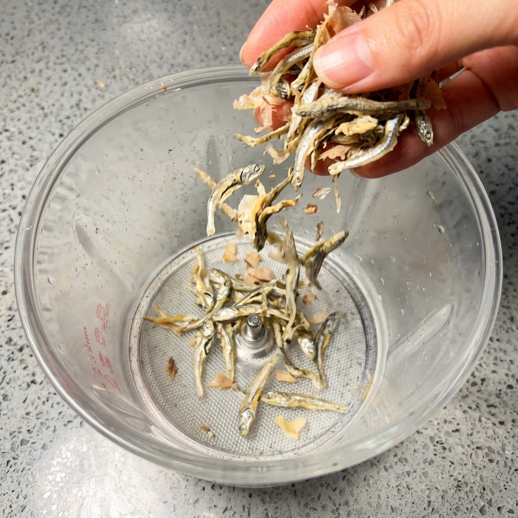 Una mano dejando caer pequeñas anchoas plateadas secas y copos de bonito marrón rosáceo en un vaso de batidora transparente.