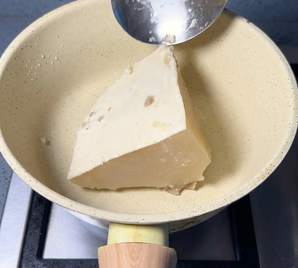 Un bloque triangular de caldo de cerdo blanco congelado en una sartén de color crema con una cuchara de metal apoyada en el borde.