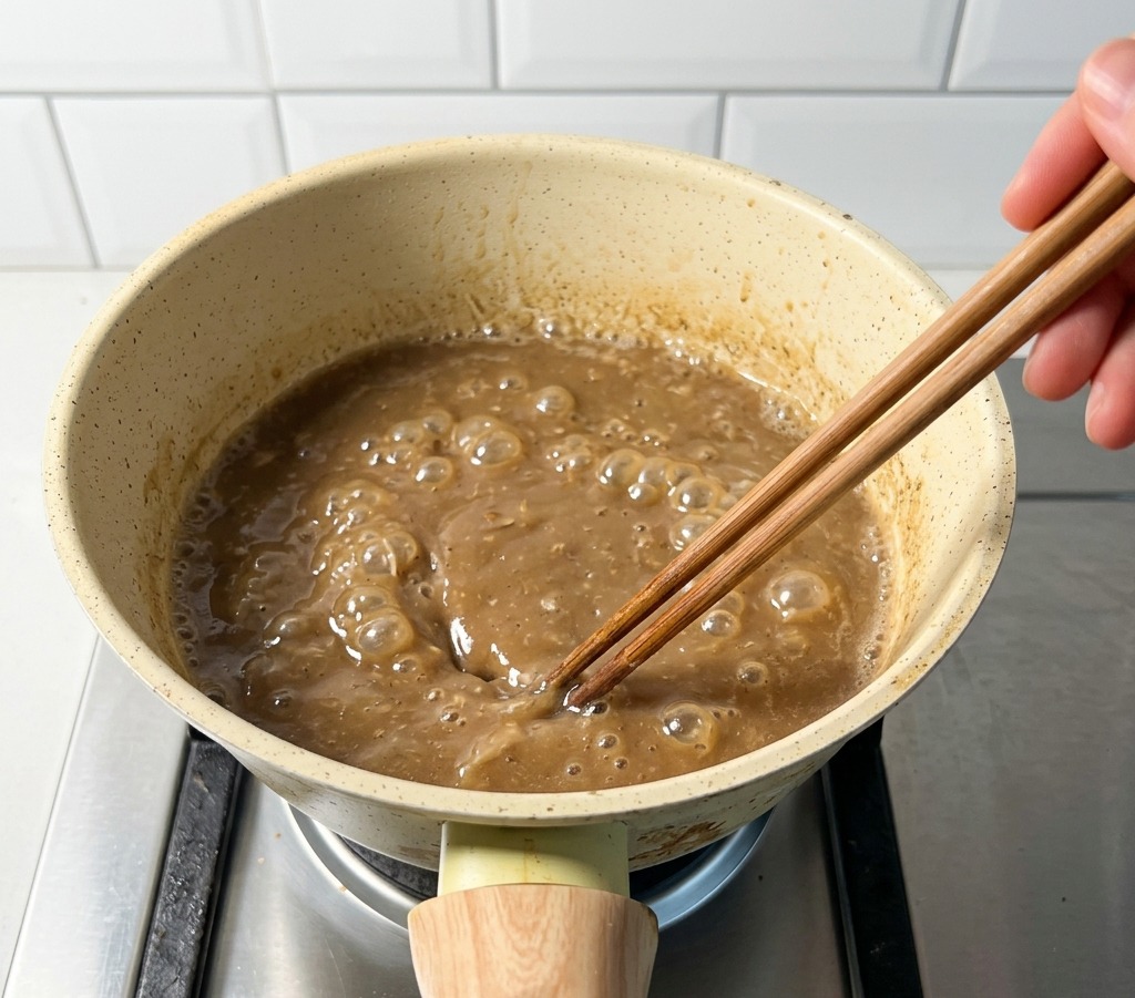 Una pequeña olla beige en un fogón de gas que contiene caldo marrón de cerdo y pescado hirviendo, removido con palillos de madera.
