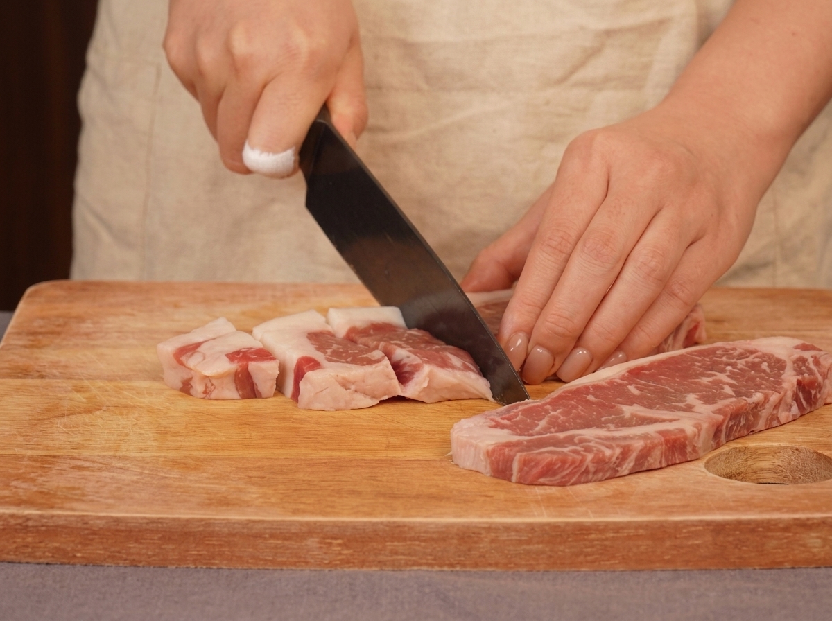 Una persona usando un cuchillo de chef para cortar carne de ternera veteada en trozos sobre una tabla de madera.