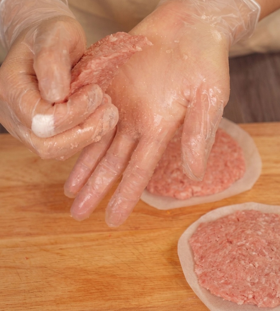 Manos con guantes presionando los bordes de una hamburguesa cruda en una tabla para crear una forma cónica.