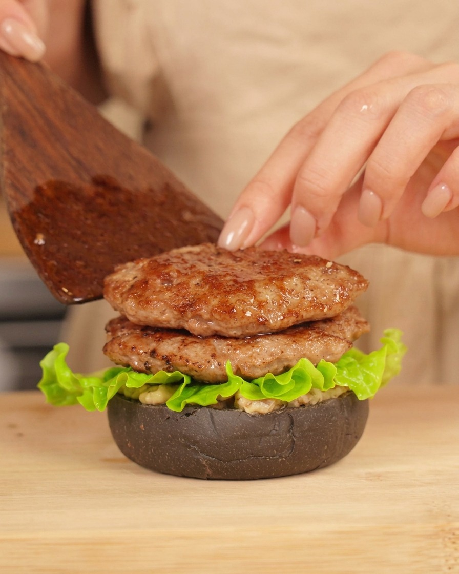 Una mano con una espátula colocando una segunda hamburguesa sobre una ya montada con lechuga fresca.