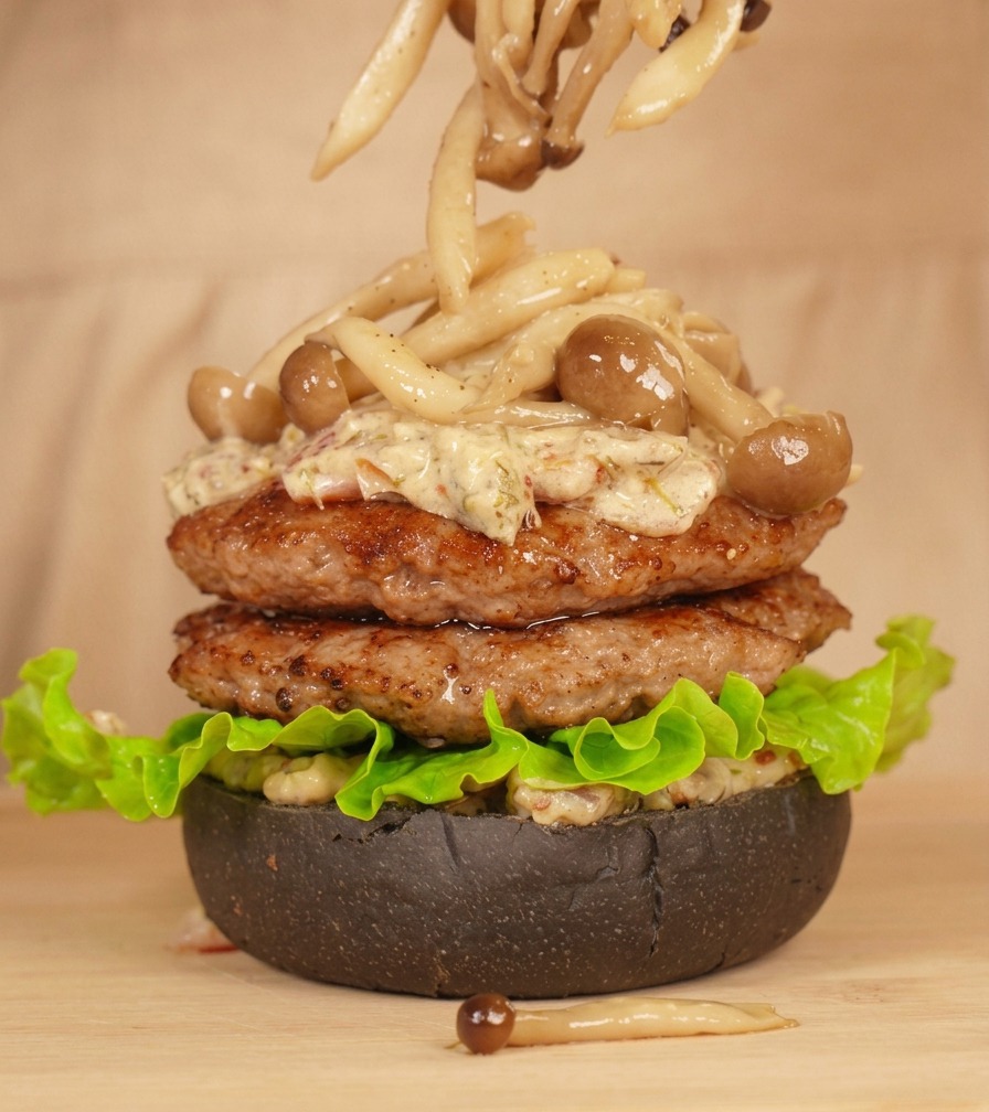 Unas pinzas colocando setas de haya sobre la torre de hamburguesas bañadas en salsa.