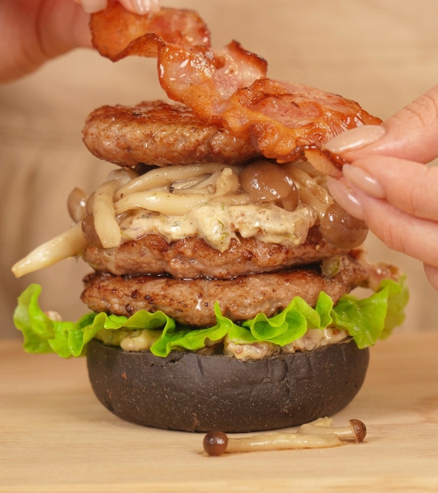 Manos colocando una tira de bacon perfectamente frito sobre una hamburguesa doble ya montada.
