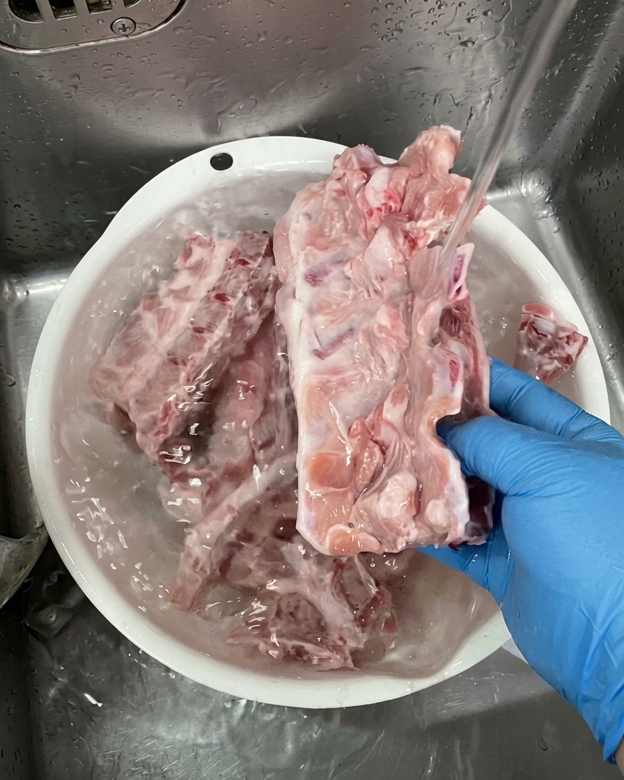 Persona con guantes azules lavando grandes huesos de cerdo crudos bajo un chorro de agua en un recipiente de plástico blanco.