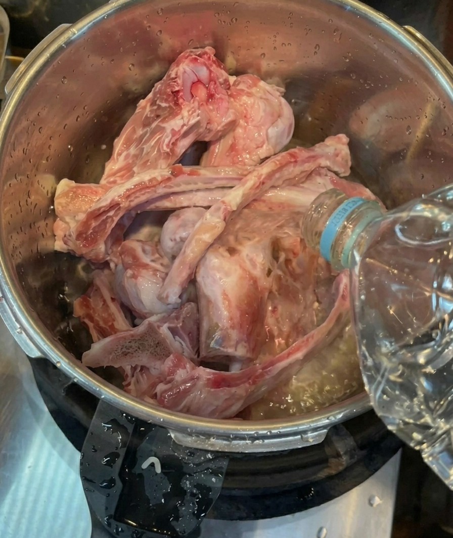 Agua embotellada vertiéndose sobre huesos de cerdo crudos y limpios dentro de una olla a presión de acero inoxidable.