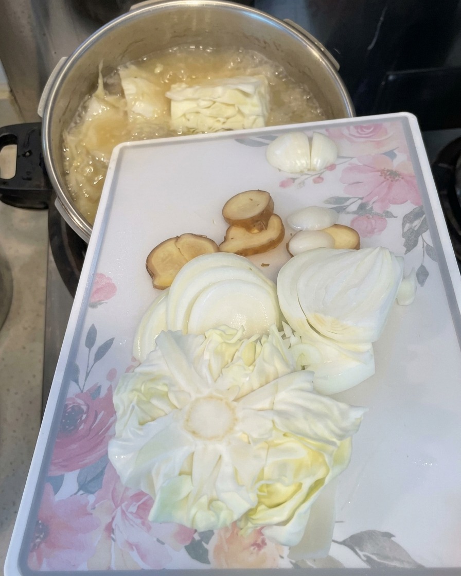 Una tabla de cortar blanca con cebollas en rodajas, jengibre y repollo inclinada para dejar caer los ingredientes en una olla hirviendo.
