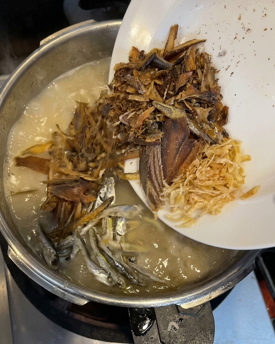 Copos de pescado seco, pequeñas sardinas plateadas y diminutos camarones secos siendo vertidos desde un plato blanco en un caldo cremoso hirviendo.