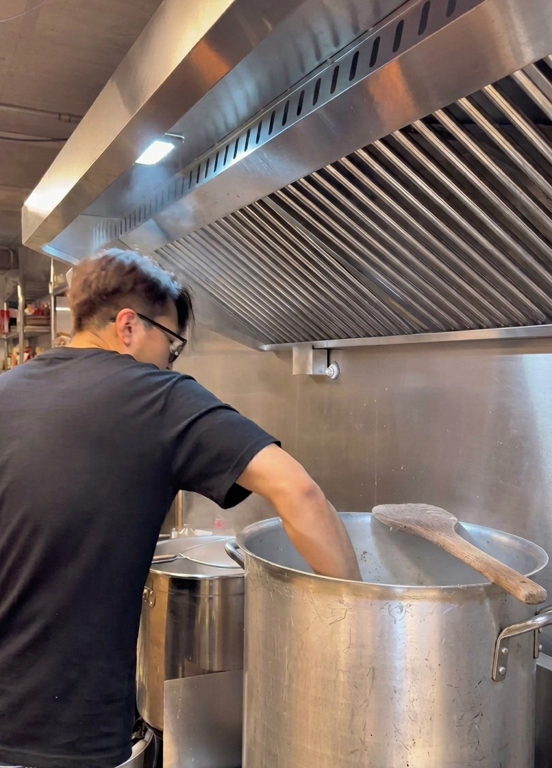 Un chef con camiseta negra se inclina sobre una gran olla de metal, colando un caldo blanco cremoso a través de un tamiz metálico.