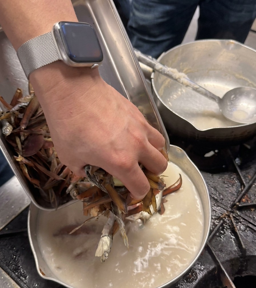 La mano de un chef añade un puñado de ingredientes secos de color marrón oscuro en una olla con caldo blanco cremoso.