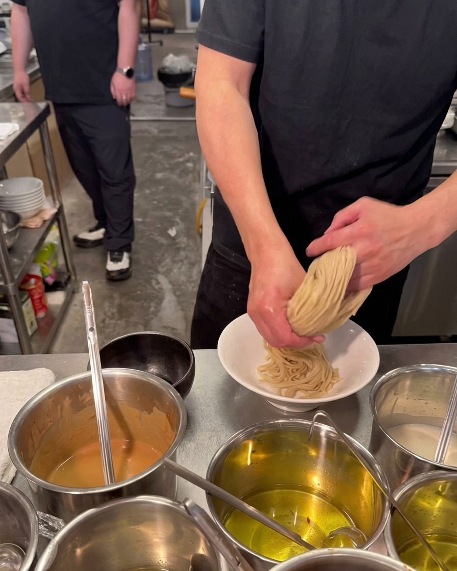 Un chef usa ambas manos para doblar y presionar un nido de fideos de ramen gruesos y pálidos en un tazón de cerámica blanca.
