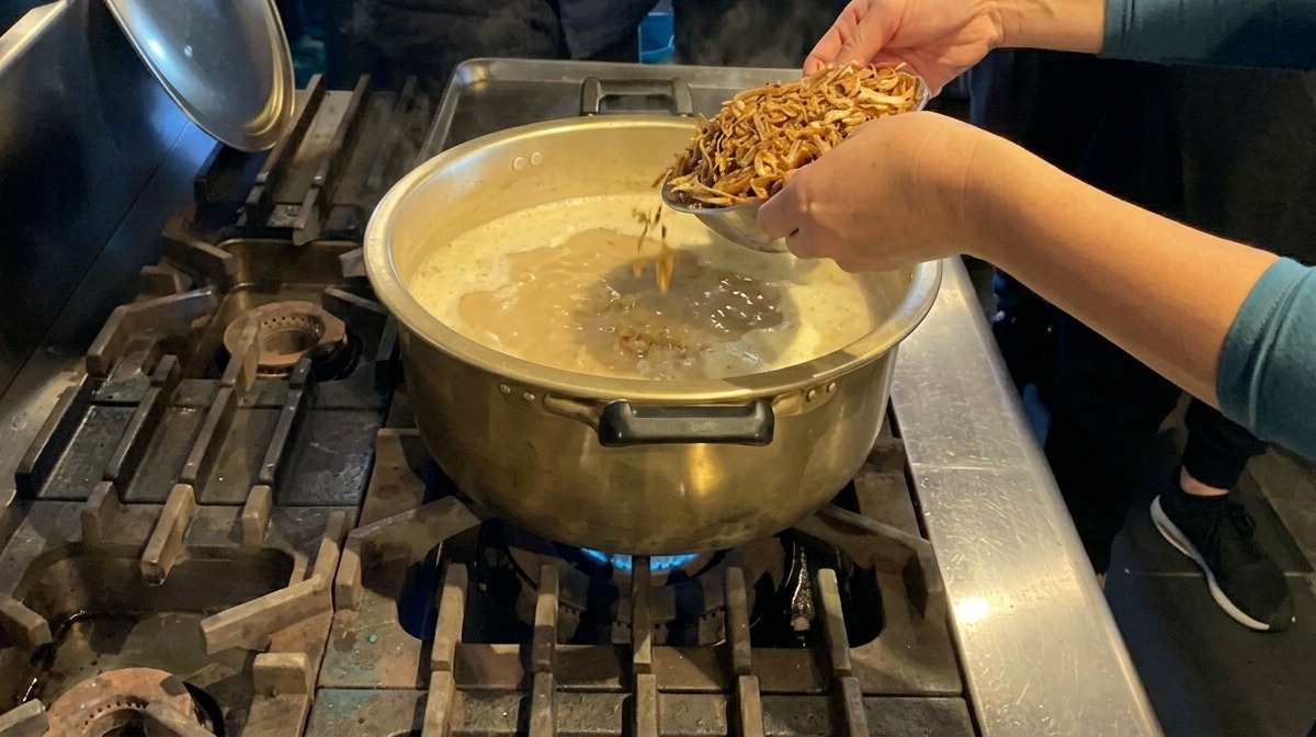 Manos agregando una montaña de anchoas secas en una olla grande de latón llena de sopa blanca y cremosa hirviendo.