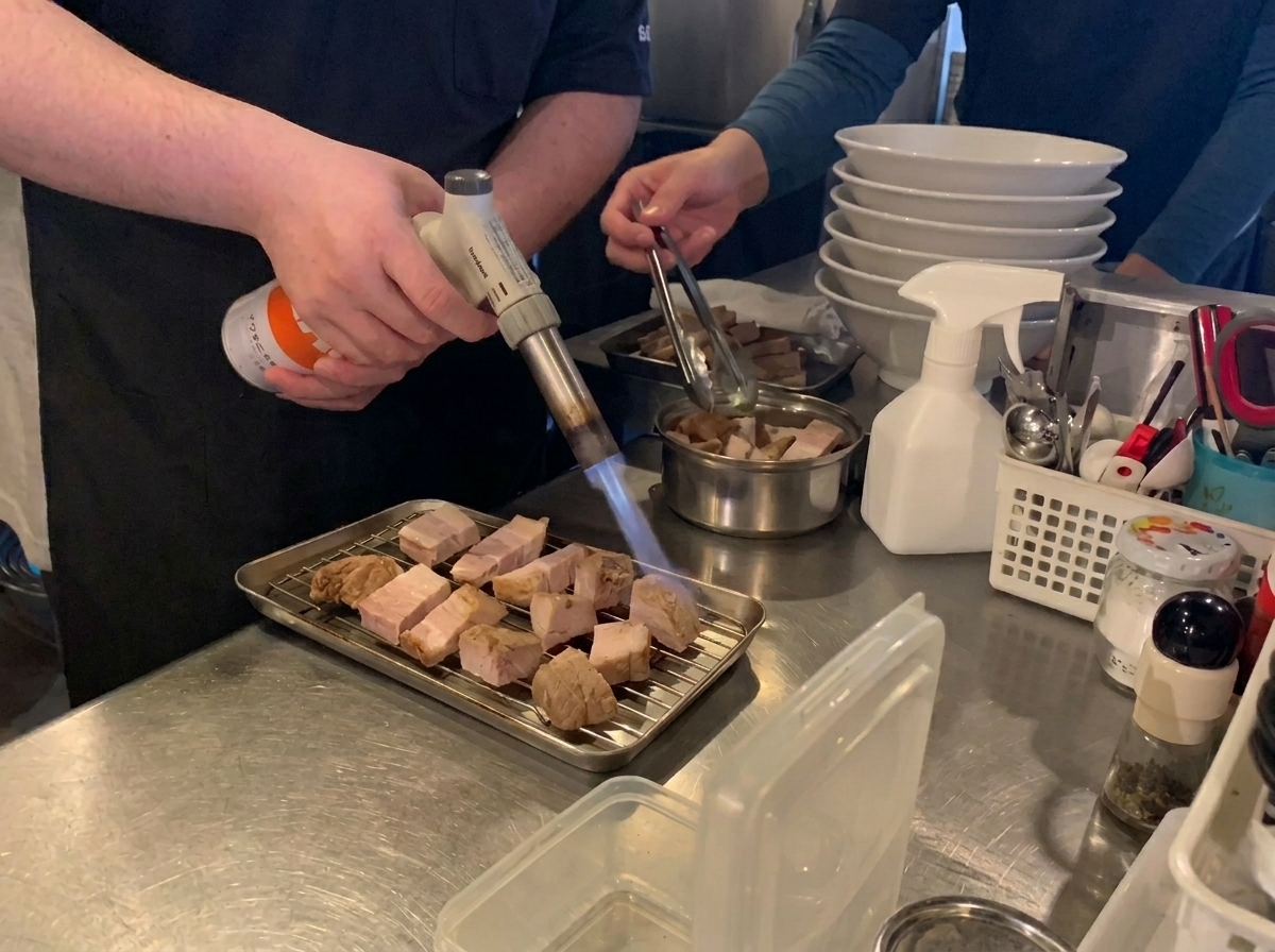 Un chef usando un soplete de butano para dorar y caramelizar rodajas de panceta de cerdo estofada en una rejilla de metal.