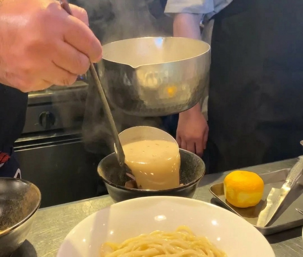 Caldo de ramen de mariscos espeso y cremoso vertiéndose desde un cucharón de metal en un bol de cerámica negra.