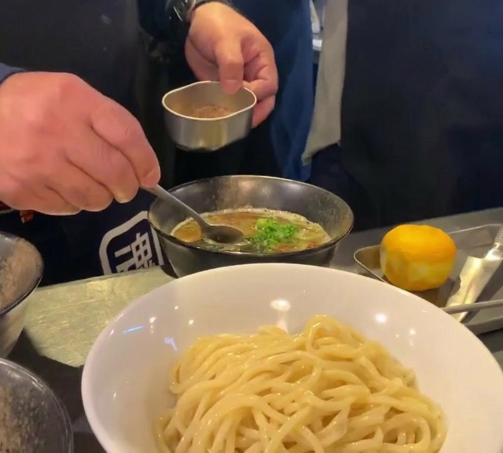 Un chef usando un pequeño recipiente de metal y una cuchara para espolvorear polvo fino de marisco marrón sobre un bol de sopa oscura y rica.
