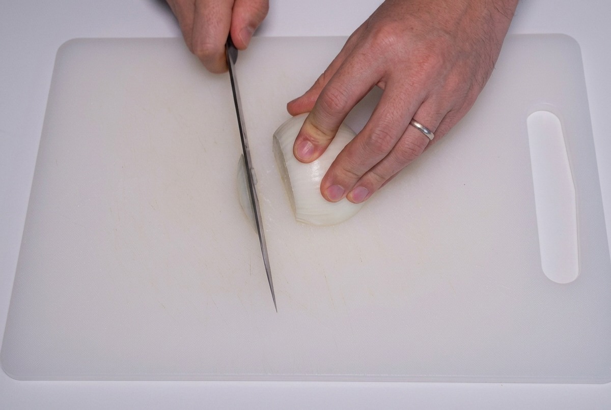 Unas manos usando un cuchillo afilado para cortar una cebolla blanca en tiras finas verticales sobre una tabla de cortar blanca.