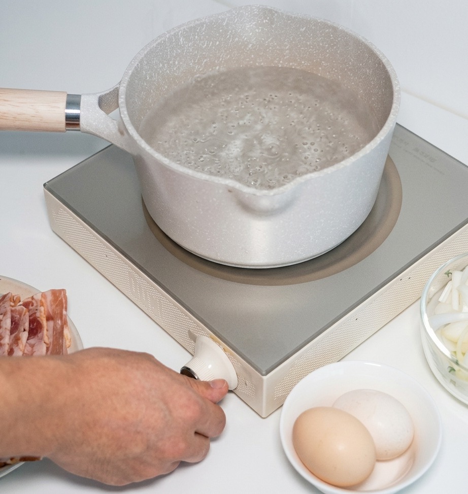 Una mano girando la perilla de control de una estufa de inducción portátil blanca con una olla de agua sobre la superficie de calentamiento.