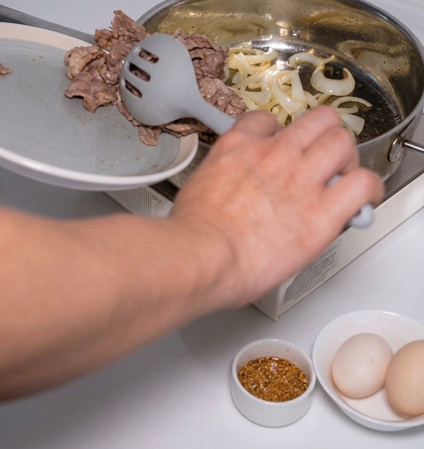 Una persona usando una espumadera gris para añadir láminas de carne cocinada a una sartén con cebollas salteadas.