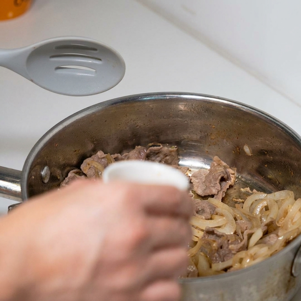 Una mano virtiendo un tazón pequeño de salsa sabrosa premezclada sobre la ternera cocinada y las cebollas laminadas en una sartén.