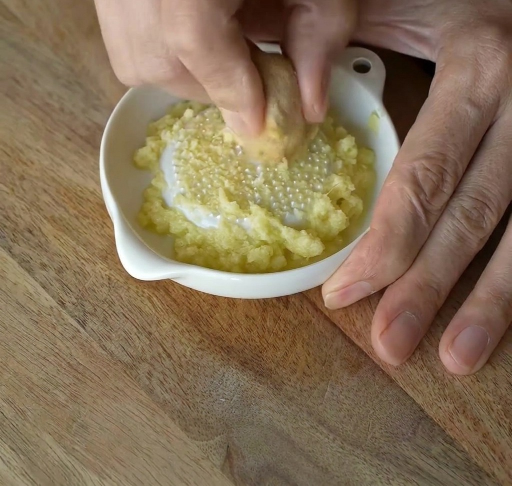 Jengibre fresco siendo frotado contra un rallador de cerámica blanco para crear una pasta fina.