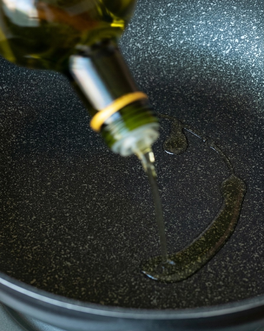 Aceite de aguacate transparente vertiéndose desde una botella de vidrio verde sobre una sartén antiadherente oscura.