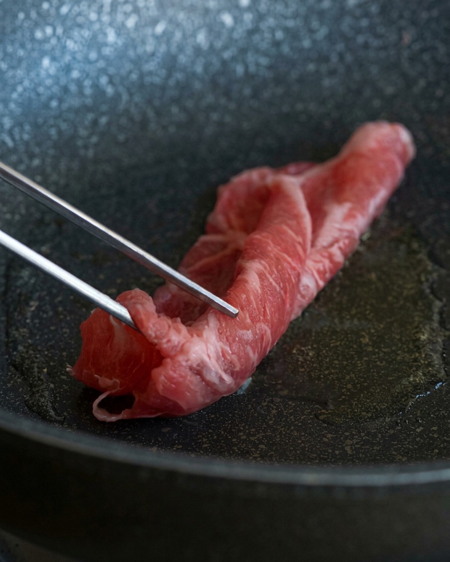 Pinzas de metal colocando una lámina delgada y marmolada de carne de res cruda en una sartén caliente con aceite brillante.