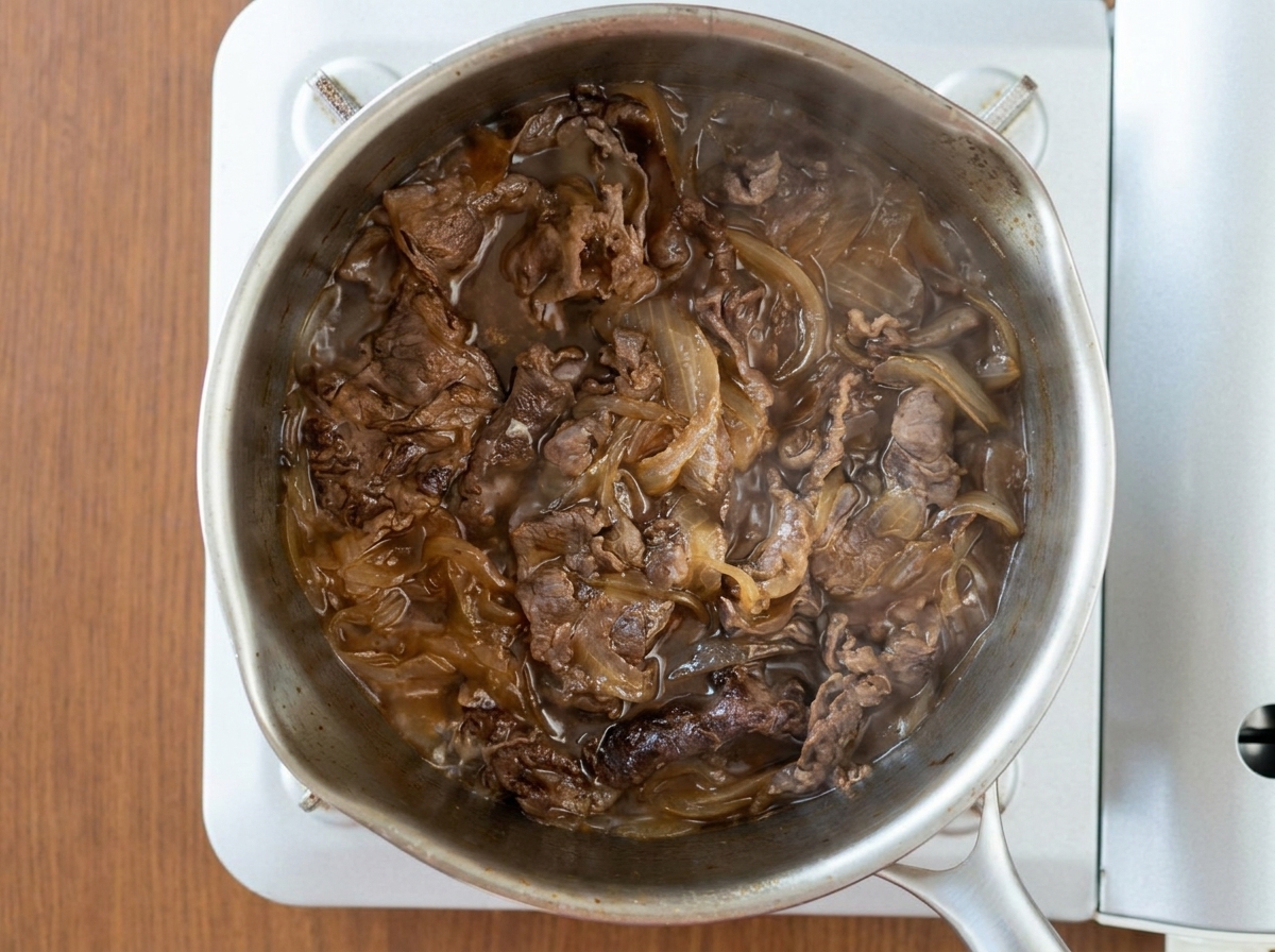 Vista aérea de láminas de carne y cebollas cocinándose juntas en una salsa oscura burbujeante dentro de una olla de acero inoxidable.