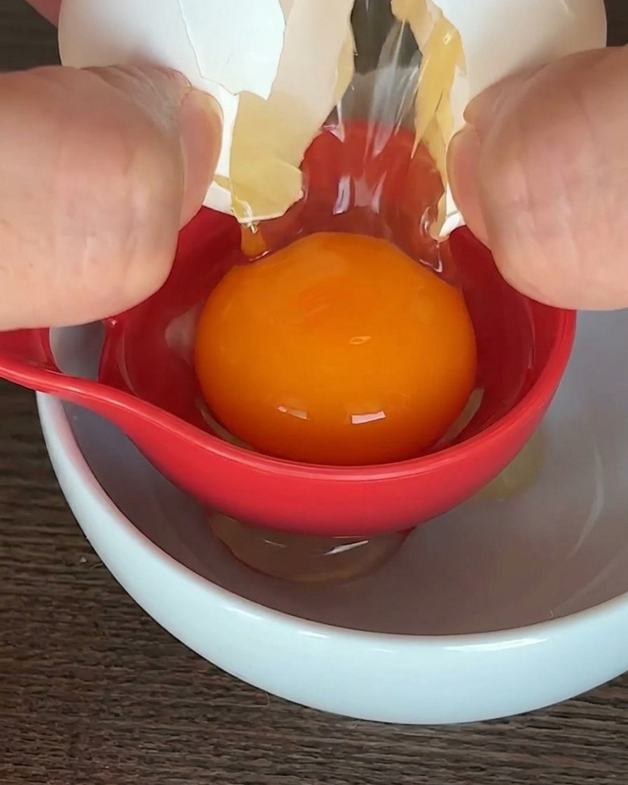 Un huevo crudo siendo separado con un utensilio rojo sobre un tazón blanco, con la yema naranja sostenida en el centro.