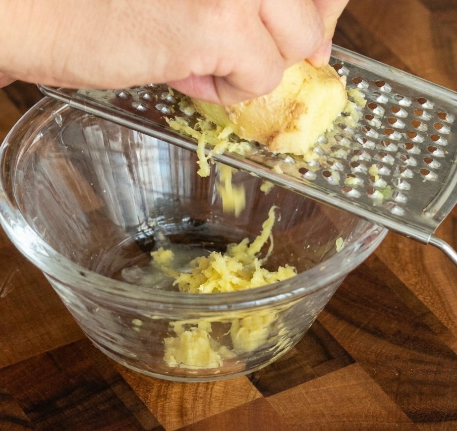 Una mano rallando un trozo de jengibre fresco en un rallador metálico, dejando caer la pasta en un pequeño cuenco de cristal.