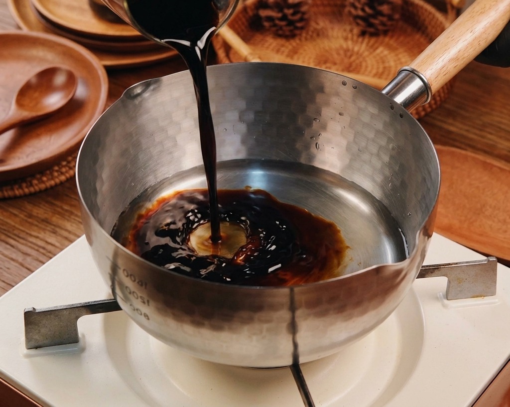 Salsa de soja oscura vertiéndose en una olla Yukihira que ya contiene agua y otros líquidos transparentes.