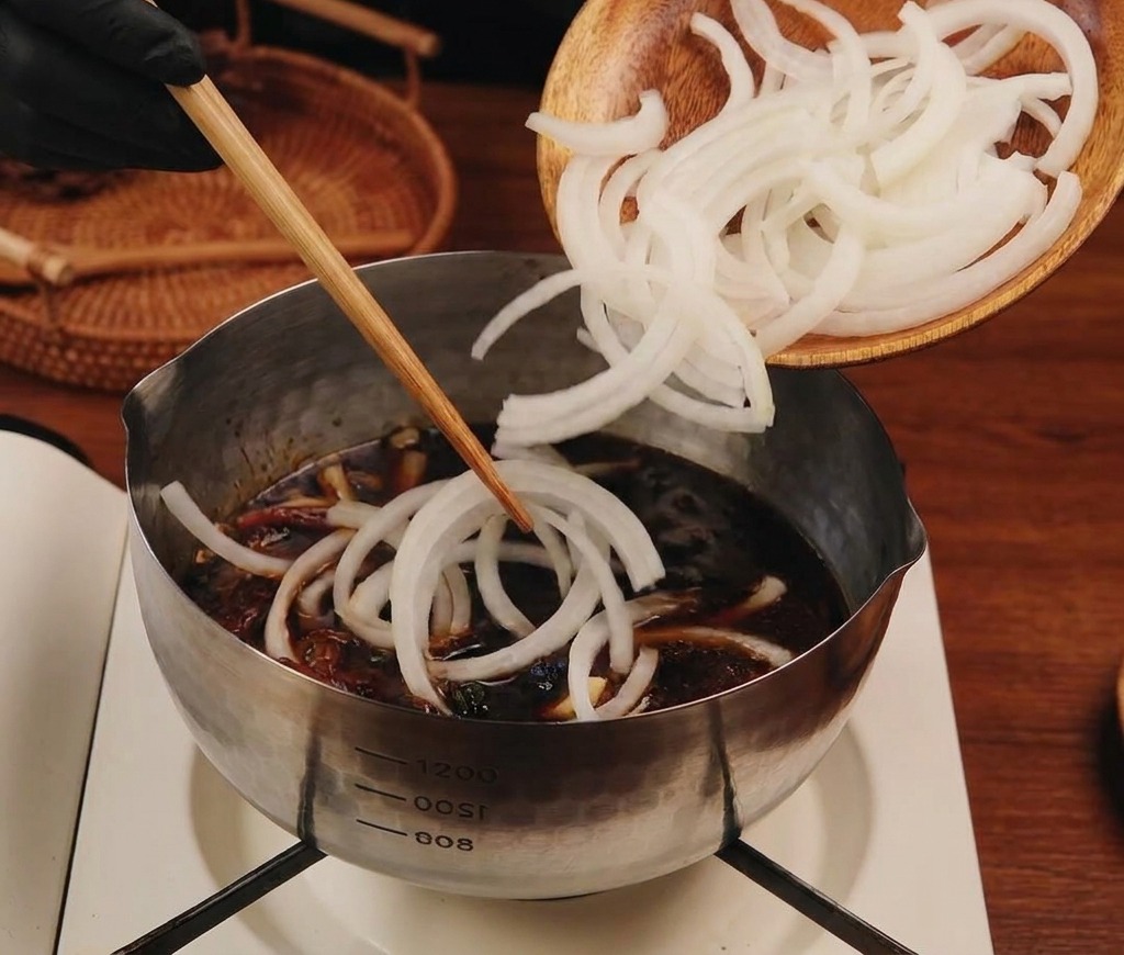 Cebollas blancas cortadas cayendo desde un plato de madera hacia una olla de acero inoxidable llena de líquido oscuro sabroso.