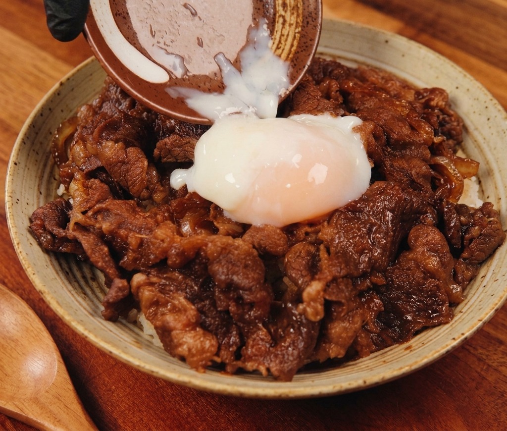Un huevo onsen poco hecho vertiéndose desde un pequeño plato de cerámica sobre un bol humeante de ternera sabrosa y arroz.