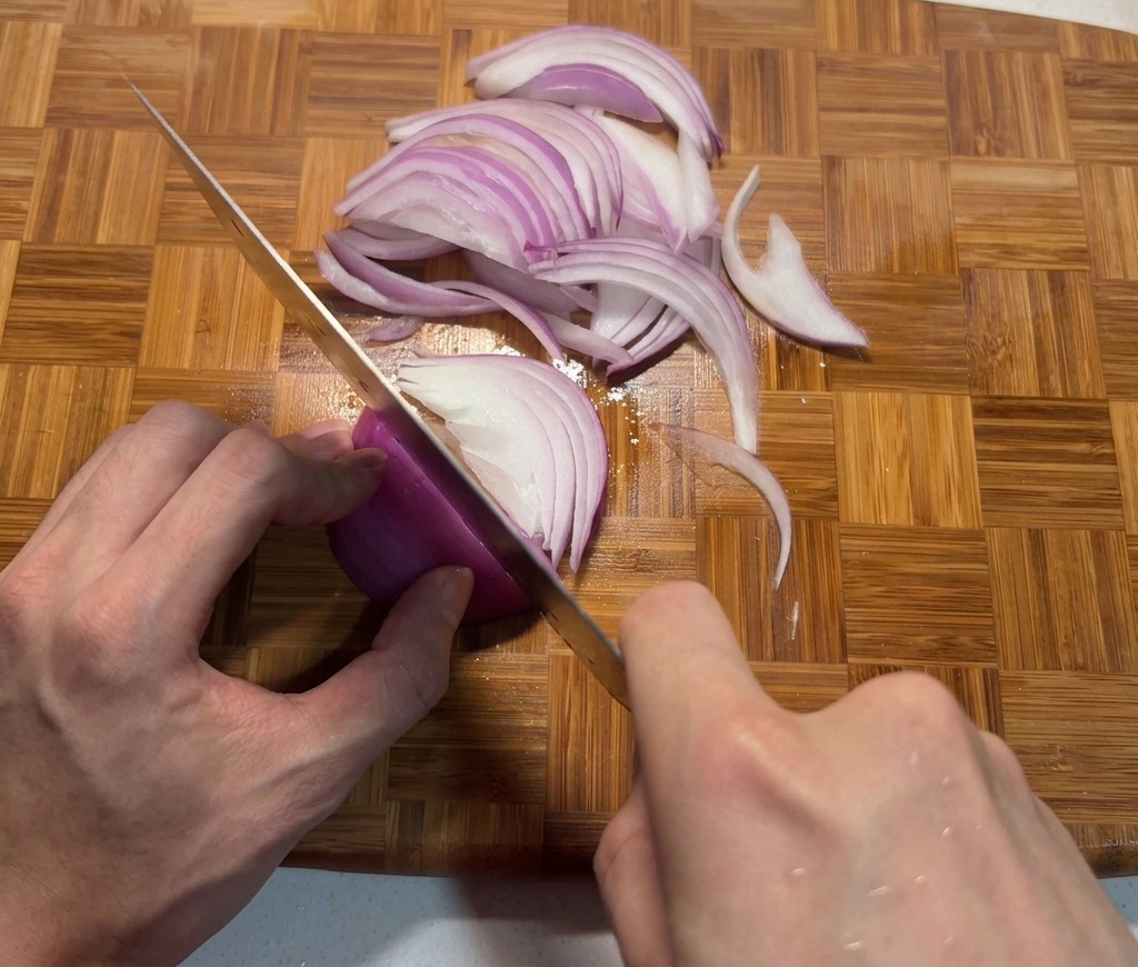 Manos cortando una cebolla morada en tiras finas sobre una tabla de cortar de bambú con un cuchillo de chef afilado.