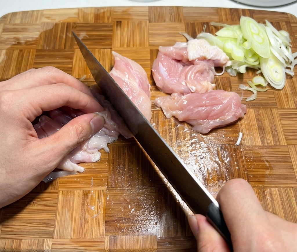 Cortando contramuslos de pollo crudo en trozos pequeños sobre una tabla de cortar de madera.