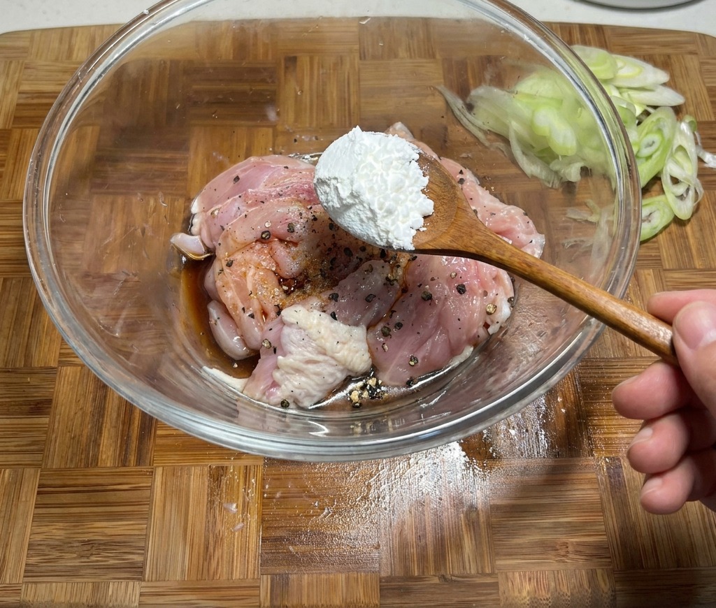 Añadiendo una cucharada de almidón de maíz a un bol de pollo crudo marinado en salsa de soja y especias.