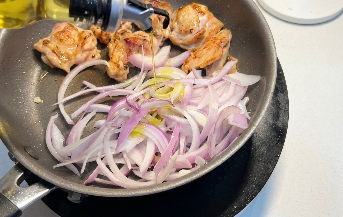 Tiras de cebolla morada añadiéndose a una sartén con contramuslos de pollo dorándose mientras se rocía un poco de aceite.