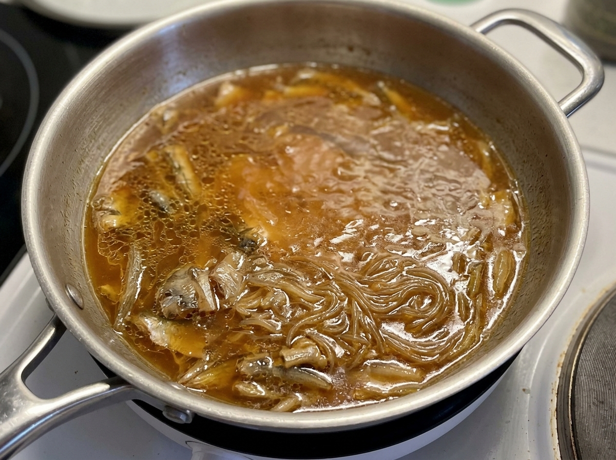 Una tapa de vidrio colocándose sobre una sartén donde el pollo y las cebollas hierven a fuego lento en una salsa marrón.