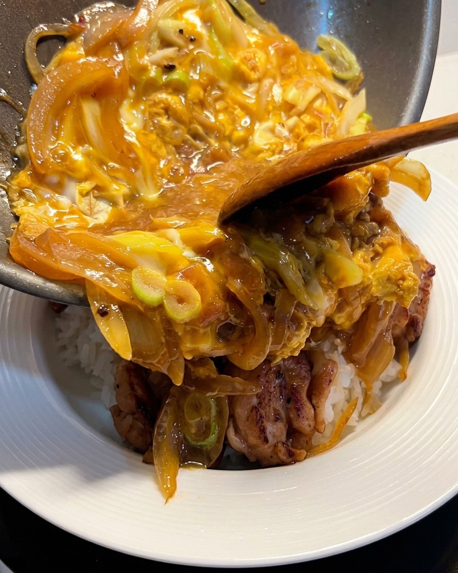 Deslizando una mezcla de huevos tiernos y cebollas desde una sartén hacia un bol de arroz blanco cubierto con pollo.