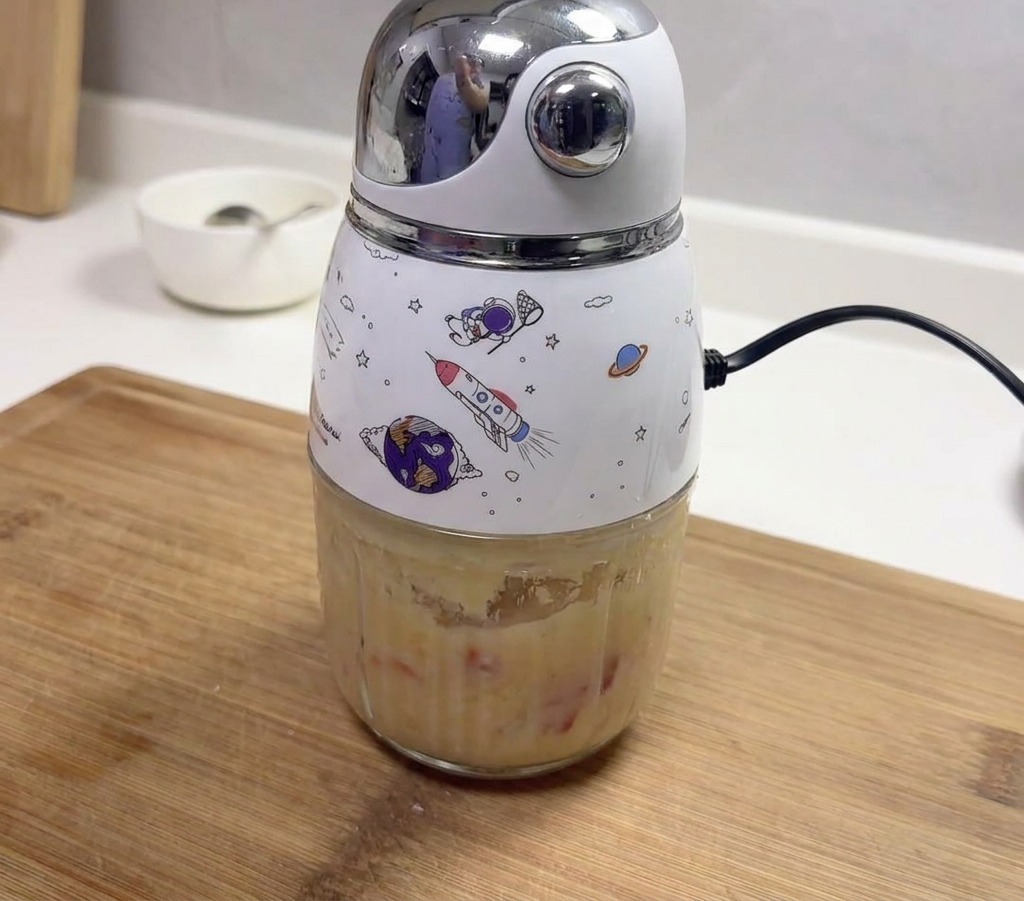 Una pequeña licuadora eléctrica decorada con motivos espaciales que contiene una pasta espesa de ajo y chiles rojos licuados.