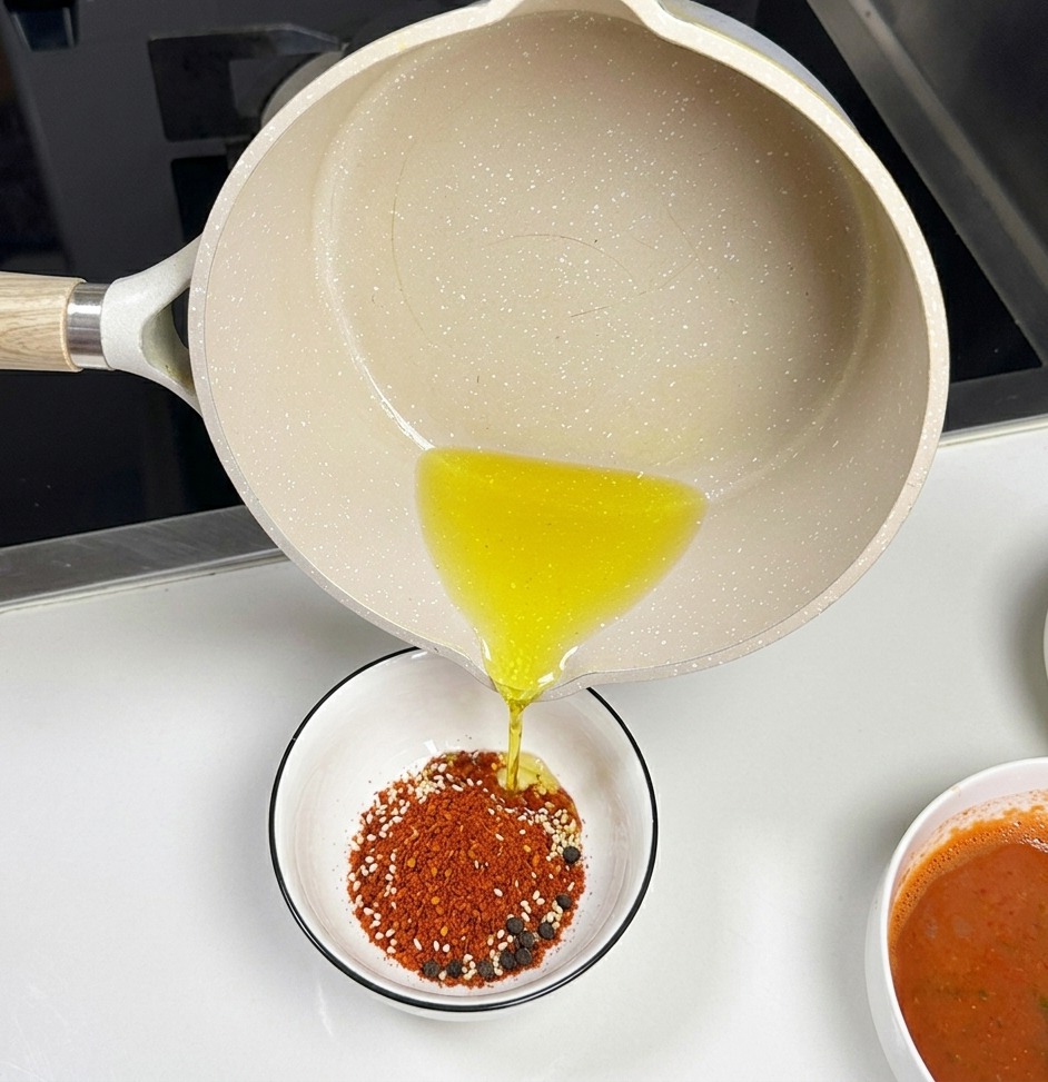 Una cacerola de cerámica de color claro vertiendo aceite caliente transparente en un pequeño tazón blanco lleno de chile rojo en polvo y semillas de sésamo blanco.
