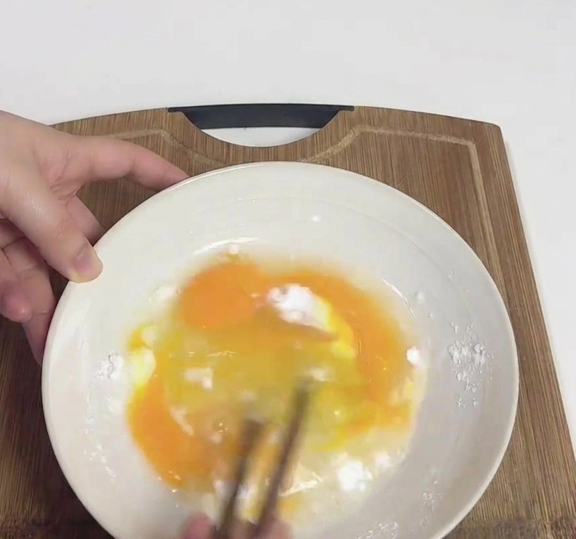 Manos batiendo huevos amarillos y fécula de patata blanca en un tazón blanco usando un par de palillos de madera.