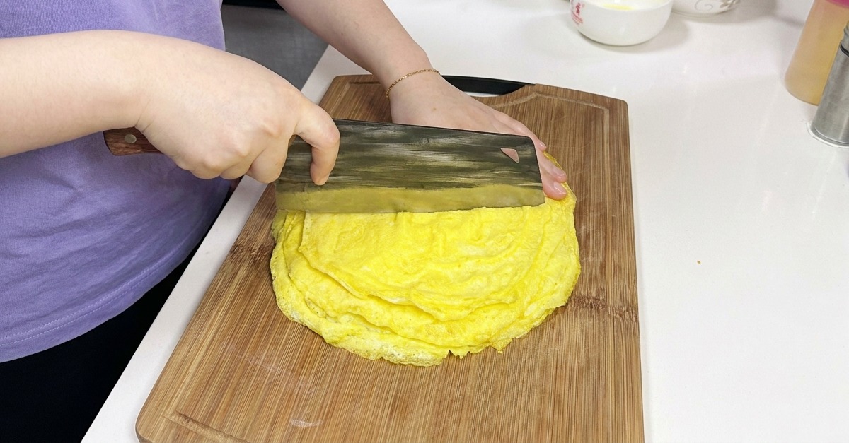 Una persona usando un gran hacha de cocina china para cortar una pila de tortillas de huevo amarillas finas y cocinadas sobre una tabla de madera.