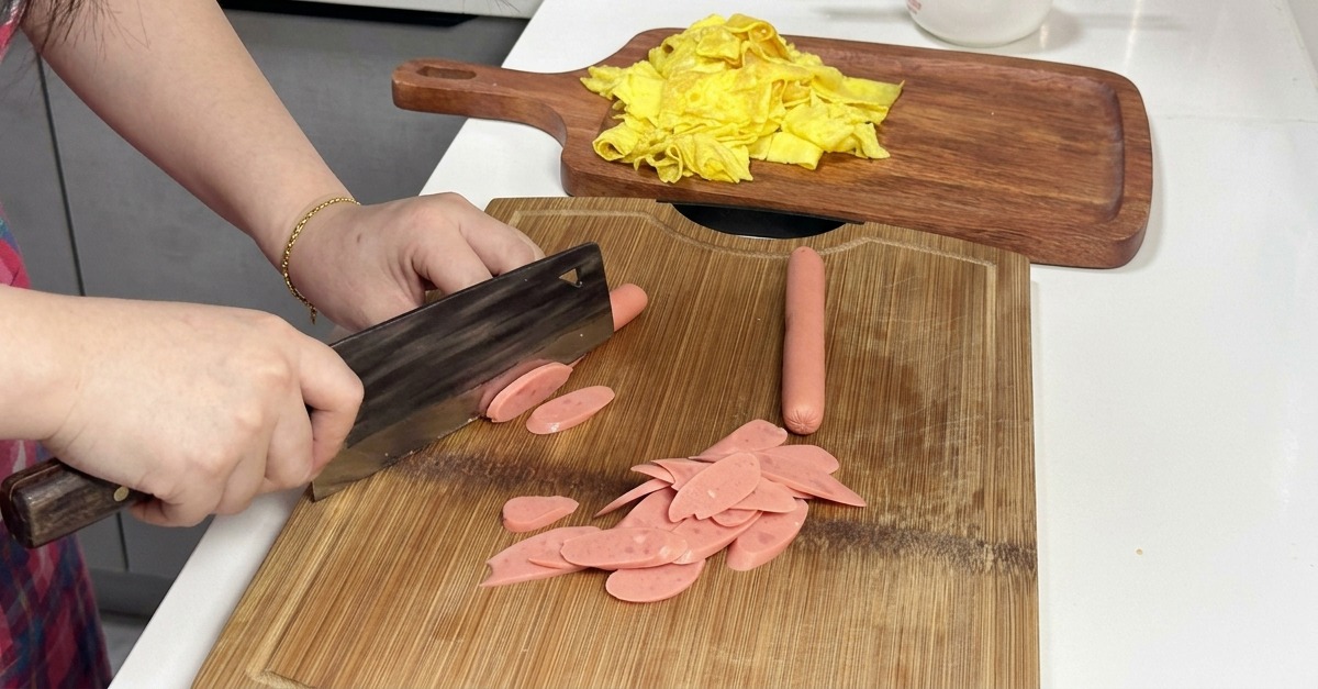 Una persona cortando salchichas rosadas diagonalmente sobre una tabla de cortar de madera con un hacha de cocina.