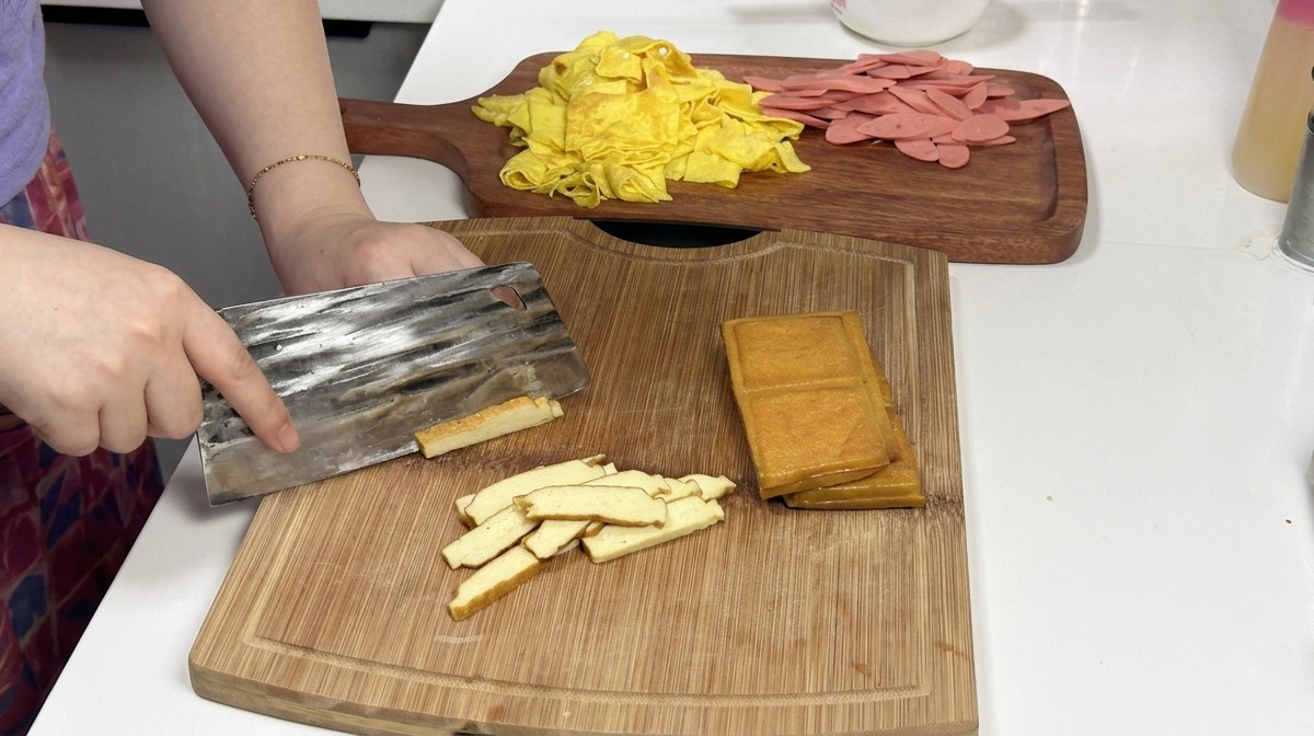 Un hacha de cocina de metal cortando una pila de bloques de tofu ahumado de color marrón dorado sobre una tabla de cortar de madera, con tiras de huevo y jamón preparadas al fondo.